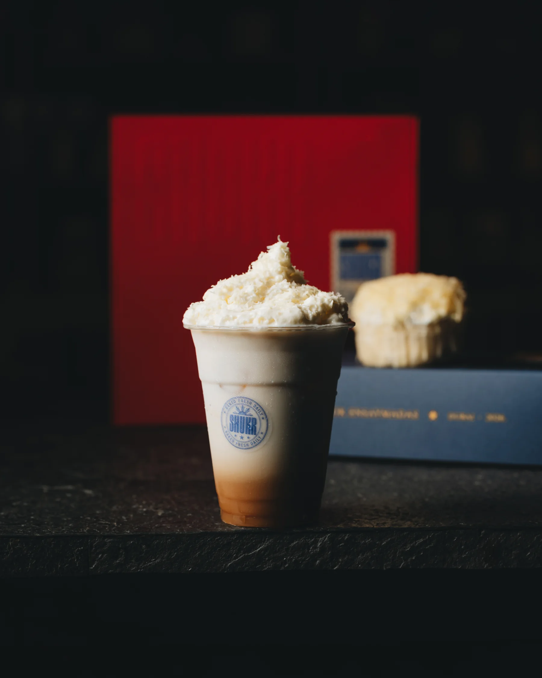 The Shukr coffee, cream on top, beside an ensaymada on the navy box
